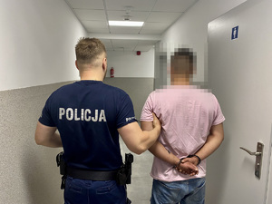 Policjant podczas doprowadzenia osoby zatrzymanej, która ma założone kajdanki na ręce trzymane z tyłu.