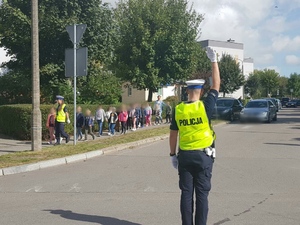Umundurowana policjantka podczas przeprowadzania dzieci przez przejście dla pieszych i policjant kierujący ruchem na skrzyżowaniu.