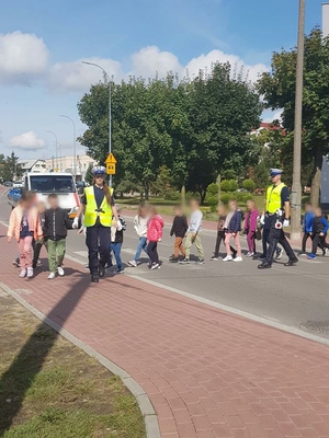 Umundurowani policjanci podczas przeprowadzania dzieci przez przejście dla pieszych.