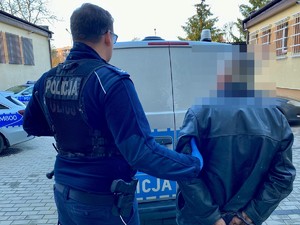 Policjant podczas doprowadzenia osoby zatrzymanej, która ma założone kajdanki na ręce trzymane z tyłu.