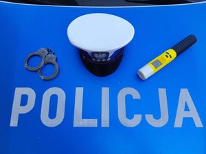 Na niebieskim tle napis &quot;Policja&quot;, czapka policyjna, kajdanki i urządzenie do badania stanu trzeźwości.